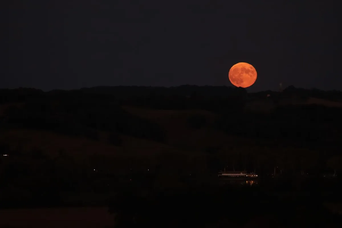 A full moon rising over a dark, tree-lined hillside at night