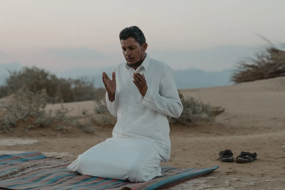 A man kneeling on a prayer mat outdoors at twilight, his hands raised in supplication