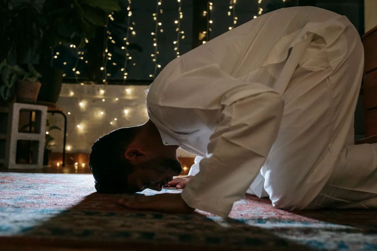 A man in prostration on a prayer rug at night, warm string lights glowing softly behind him